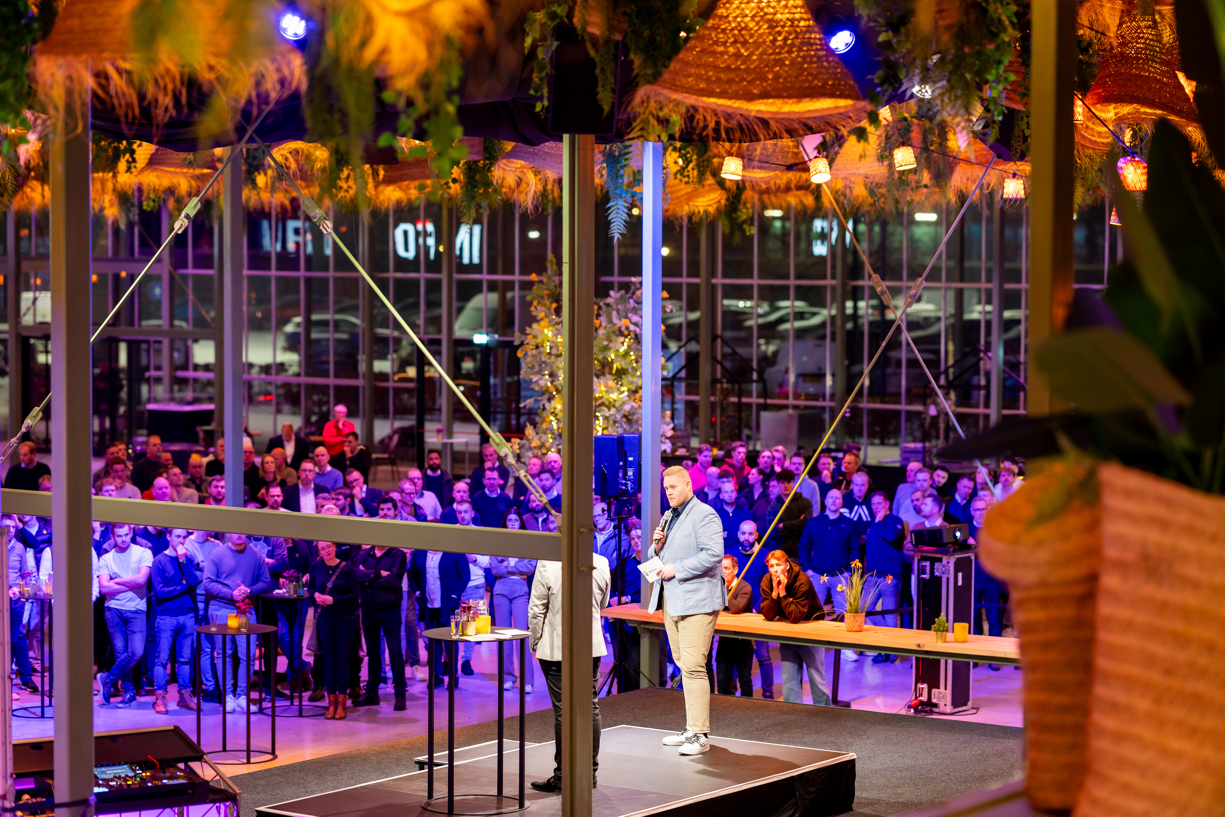 Spreker op centraal podium tijdens interactief congres, omringd door bezoekers in een informele setting.